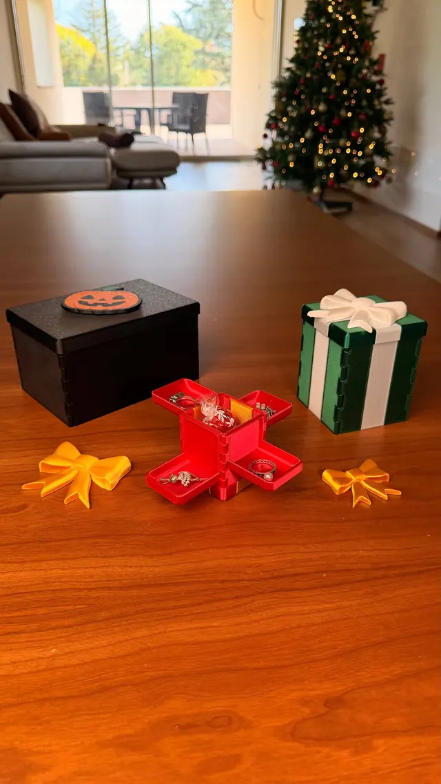 A red Gift Reveal open on the table, with rings and necklaces on its shelves, between a black box and a green one with a white ribbon